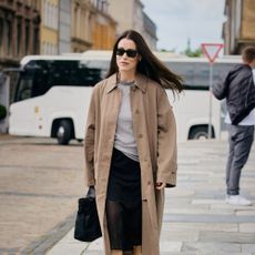 Woman wears trench coat black skirt, heels, and black handbag. 