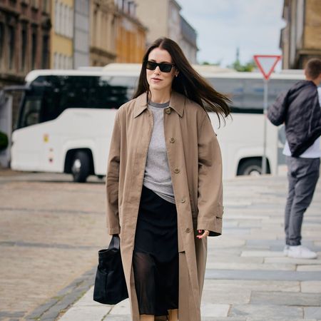 Woman wears trench coat black skirt, heels, and black handbag.