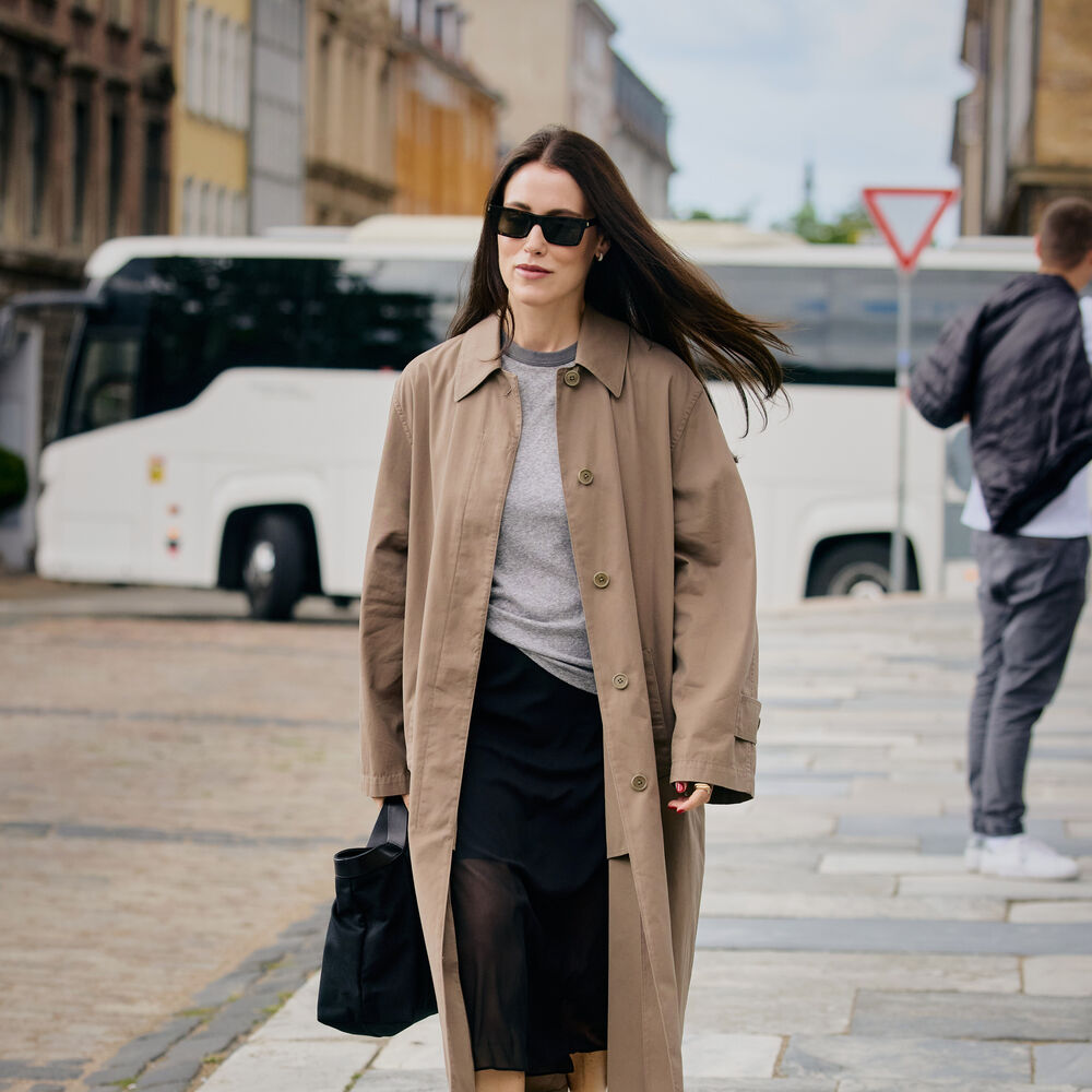 Woman wears trench coat black skirt, heels, and black handbag. 