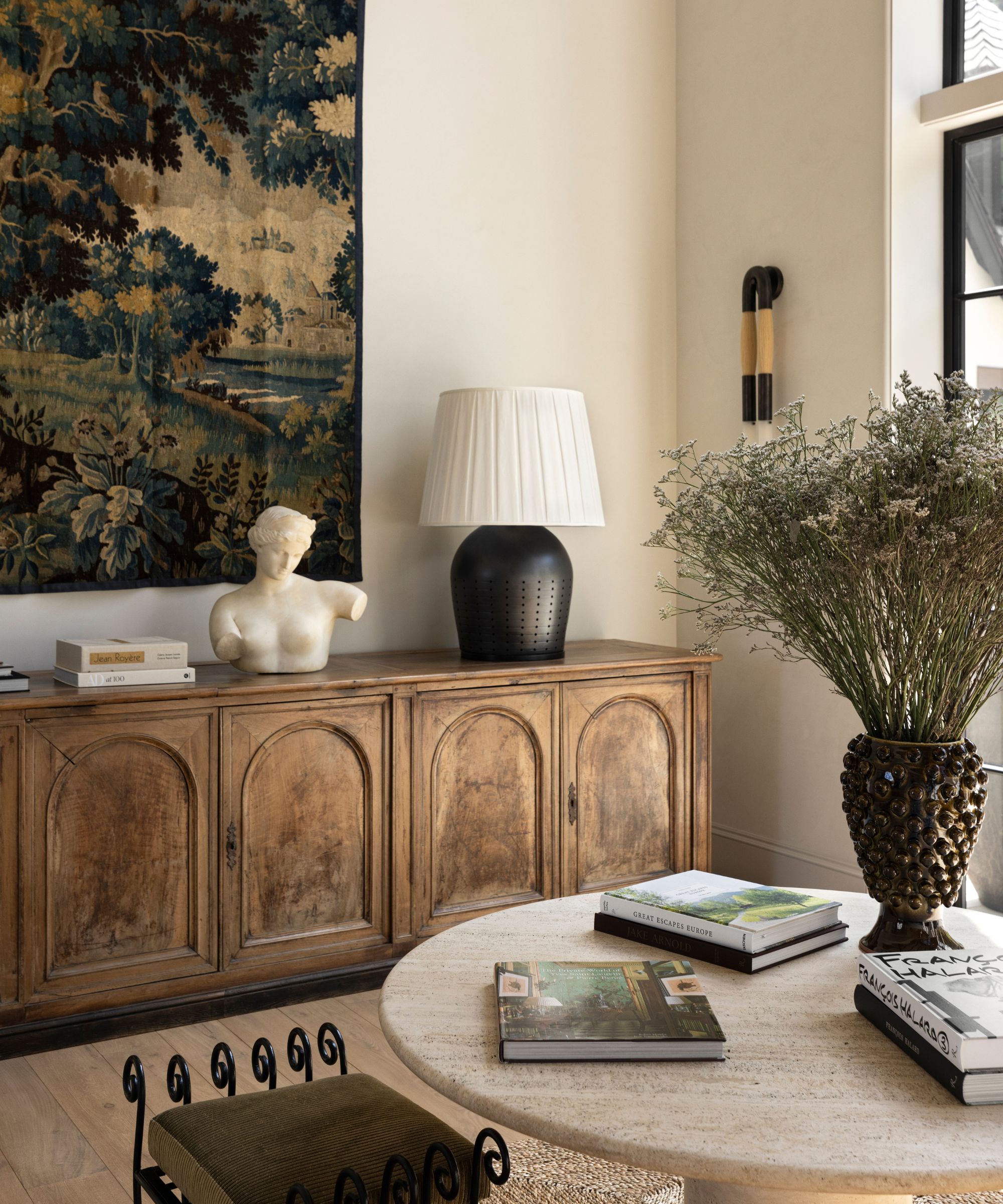 Entryway with round marble table, vintage books, wooden console with marble bust and lamp and large tapestry on wall
