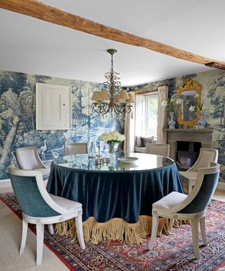 beamed blue dining room with scenic wallpaper fringed blue velvet tablecloth and Gustavian style dining chairs