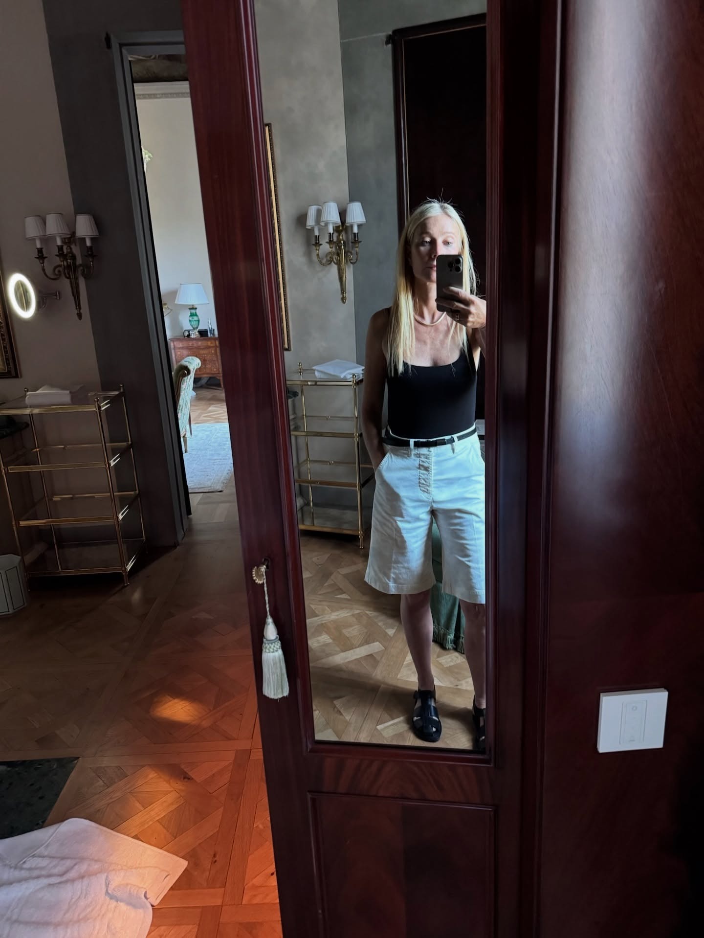 Celebrity Gwyneth Paltrow wears white bermuda shorts, a black fisherman sandal, a black belt and a black bodysuit. She is standing, posing for a selfie in a hotel room in a recent trip to Italy. This photo was taken from her Instagram recently.