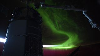 the dark, nearly silhouetted shape of a cylindrical spacecraft rises from the left, briefly reflecting the slice of white light curved from the horizon of Earth from space, seen above. a robotic arm reaches to the mass from the right, as behind a swath of green aurora smears across the dark face of the planet.