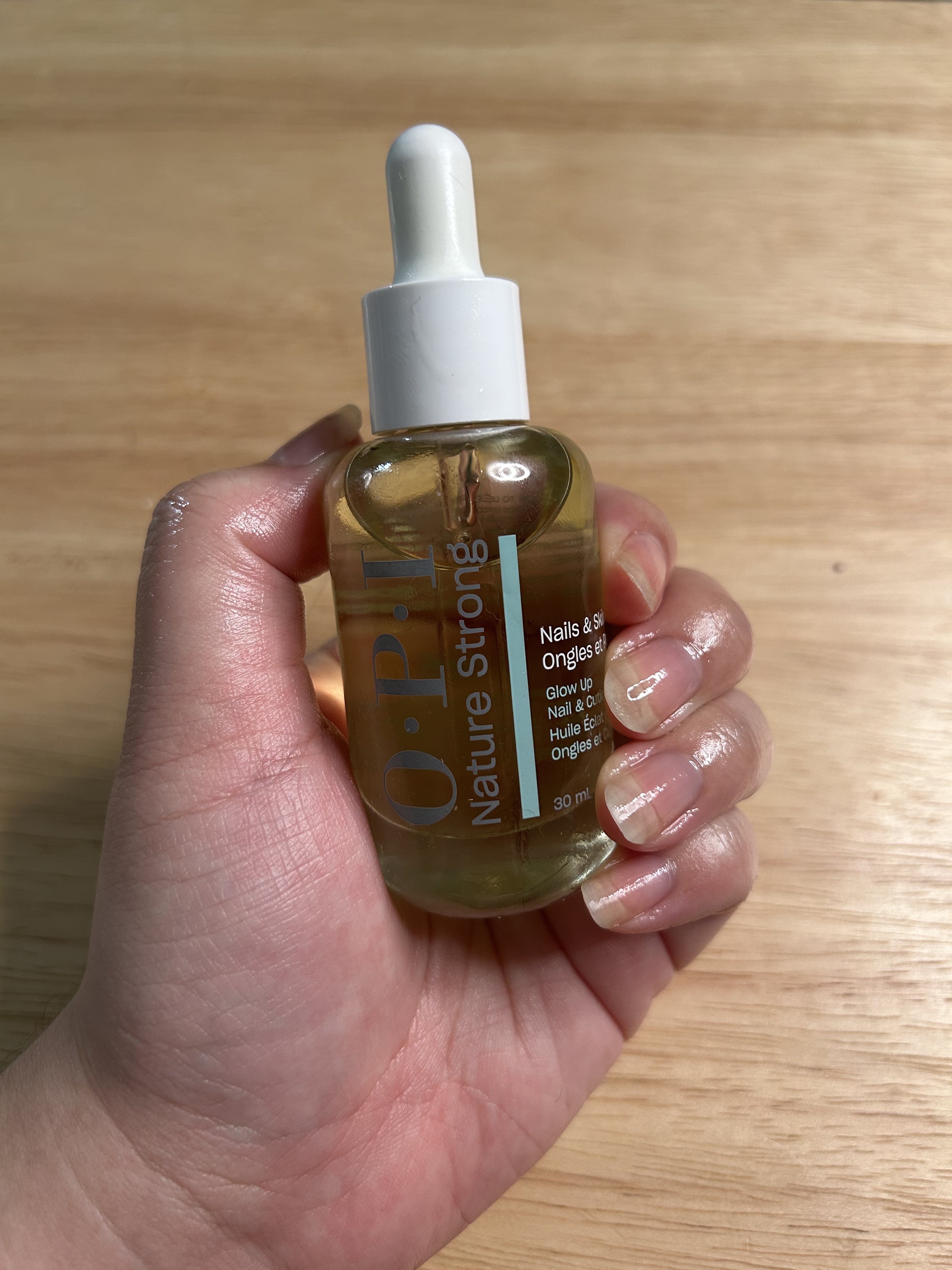 A woman's hand and nails after applying cuticle oil with a wood background holding a yellow, blue, and white bottle dropper.