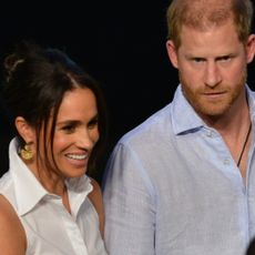 Meghan Markle wears a white, sleeveless shirt and gold earrings and wears her hair in a sleek updo, while Prince Harry wears a light blue shirt