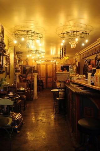 A golden-lit cafe and antiques shop houses retro-fueled interiors including Art Deco chairs and bar, plenty of wood details, and collectibles, as well as a man hurrying behind its counter.