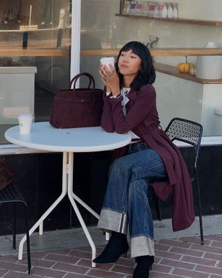 A woman wears a burgundy coat, cuffed blue jeans, and black suede ankle boots.