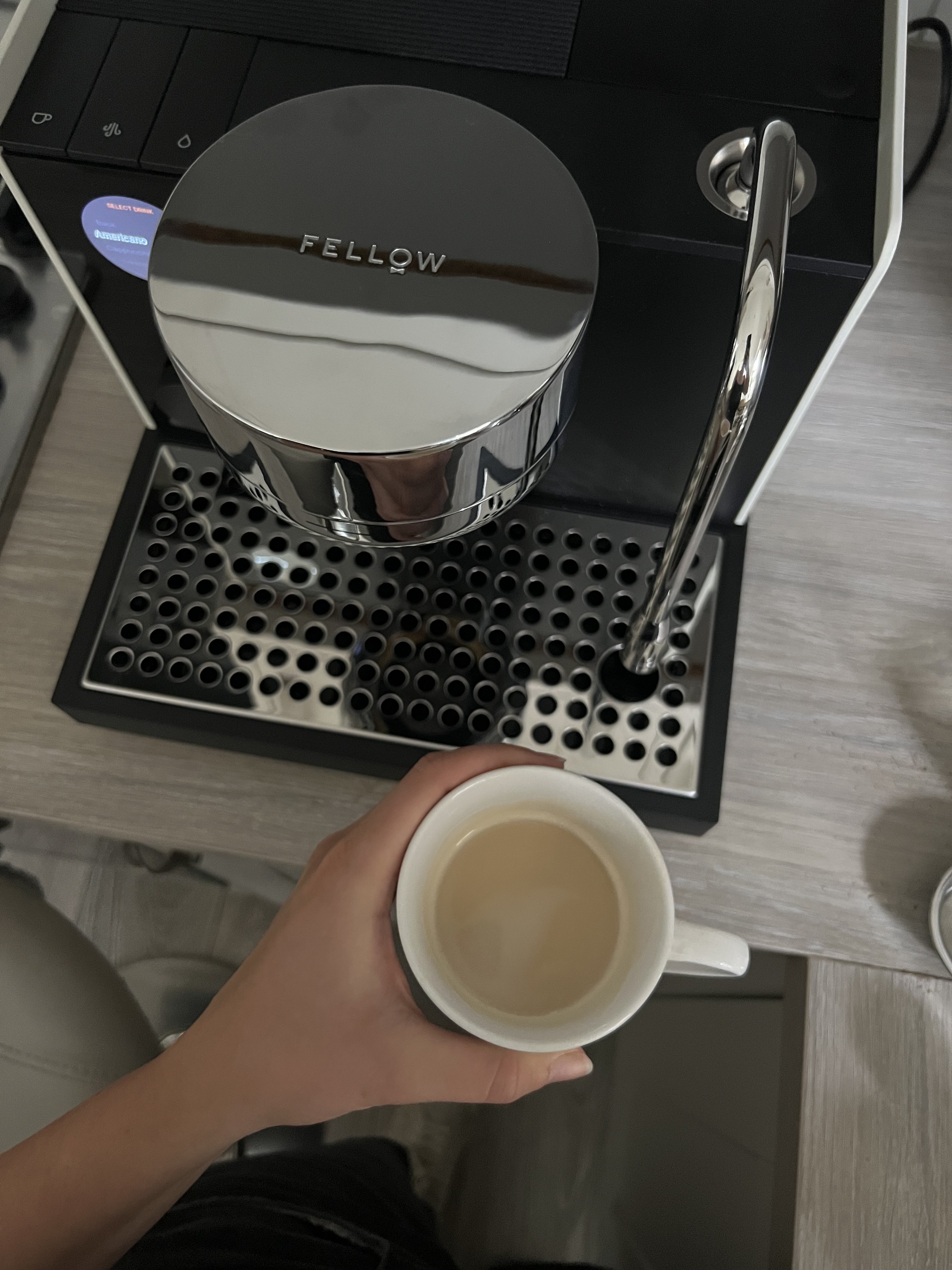 A hand holding a white mug with an espresso shot in it, in front of a black Fellow series 1 coffee machine.