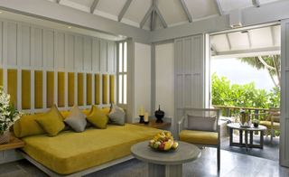 Surin hotel room with white wood panelling and exposed beams, bed with yellow bedding and cushions, round table with fruit and small balcony