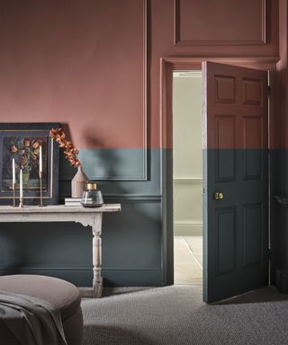 Two-toned painted bedroom with dark red-pink on upper walls, dark gray-blue on lower half, slim paneling on walls, cream carpet, rustic white wooden console table decorated with vases, artwork, candles and books