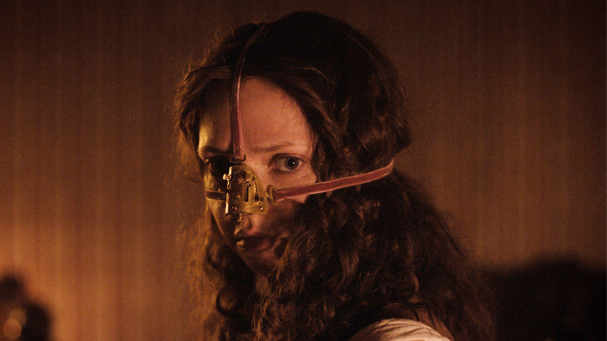 A girl, Elvira, stares at the audience. A metal nose covering is tied to her face.