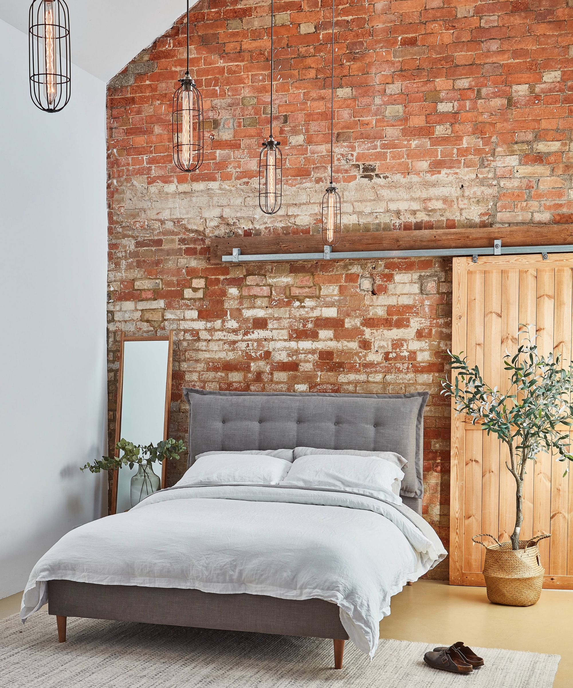 Grey upholstered bed against an exposed brick wall, with cage pendant lights and a sliding wood door.
