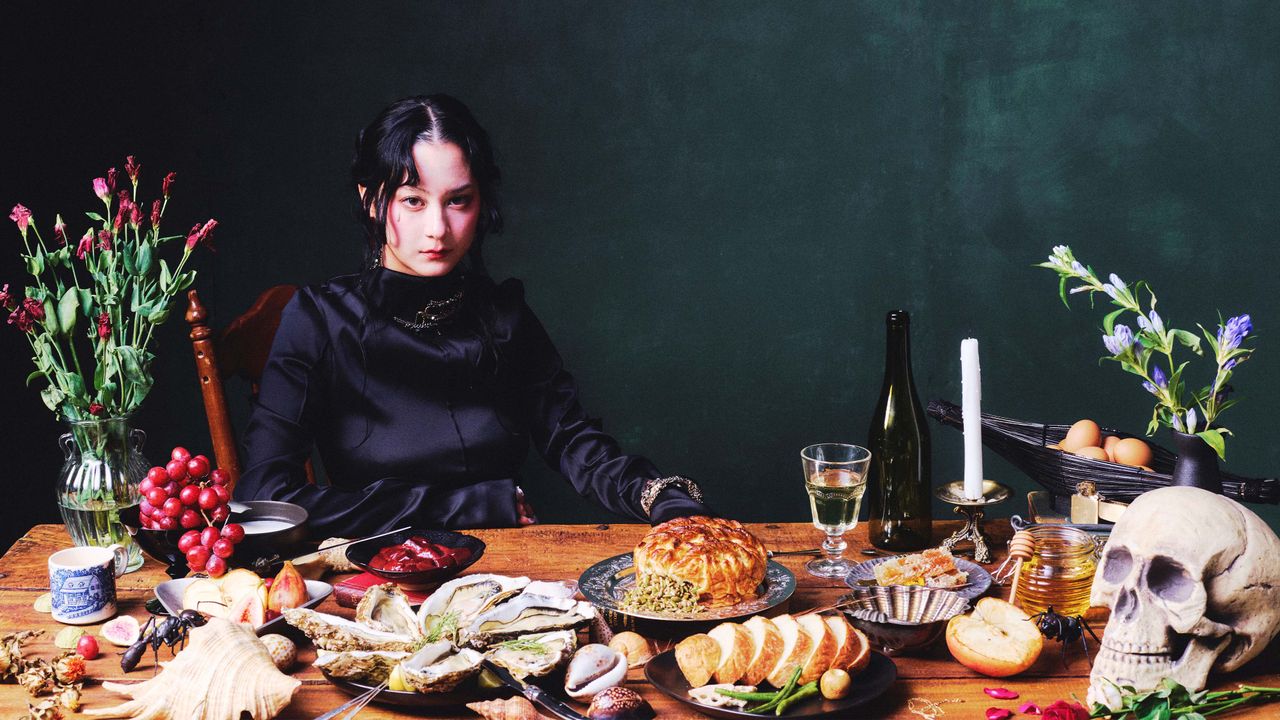  michelle zauner of japanese breakfast wears a black silk dress and sits at a table with lots of food 