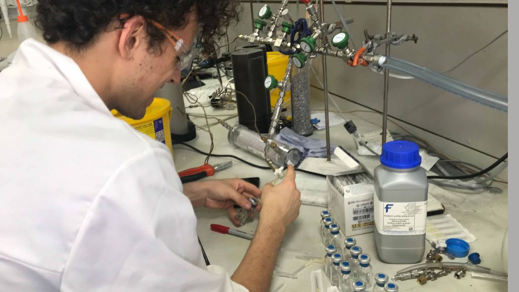 Scientist in a lab coat is pictured injecting something into a small glass vial on a lab bench