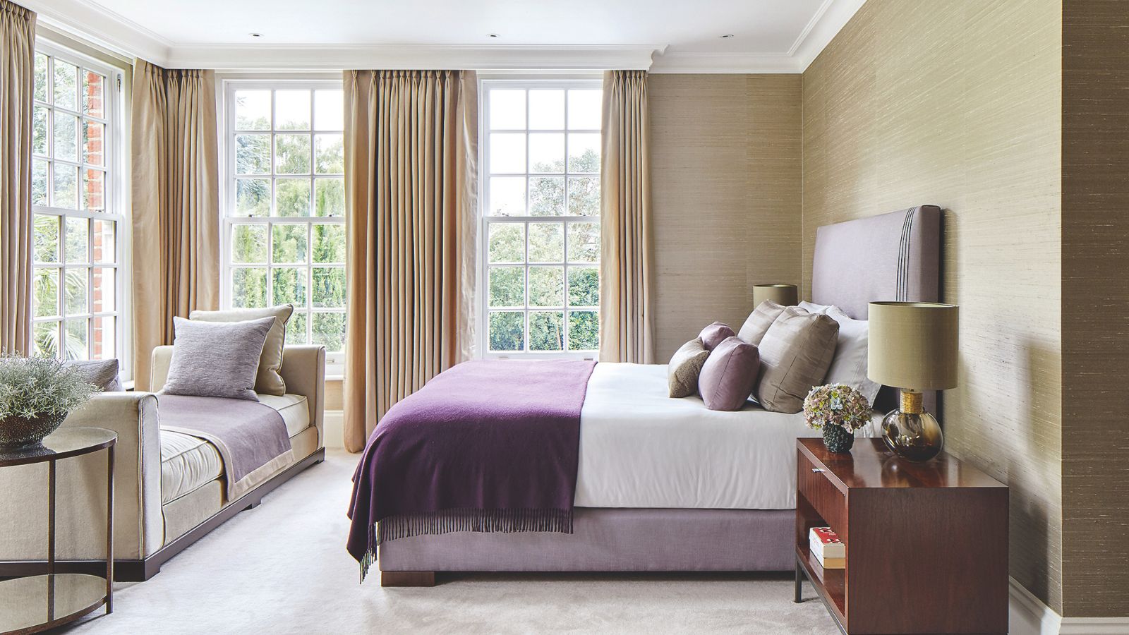 Main bedroom with grasscloth wallpaper and textured curtains, purple and white bedlinen, a chaise longue and wooden bedside table.
