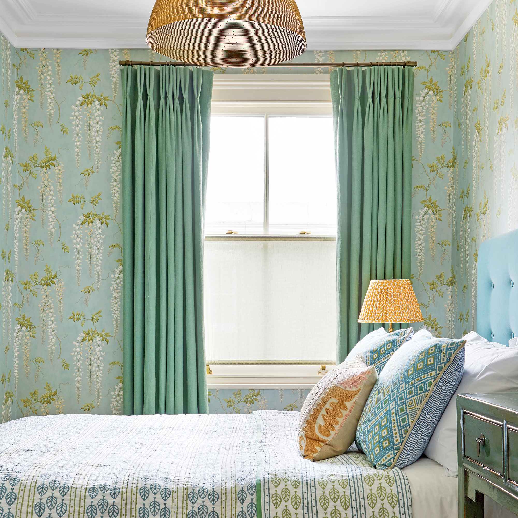 Green bedroom with wisteria wallpaper, green patterned bedding and rattan lampshade