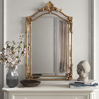 an elaborate gold mirror sits atop a fireplace with a ceramic bust and vase filled with flowers