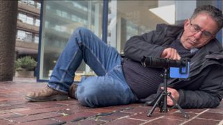 Shot of a mini tripod mounted iPhone standing on the floor of a tiled walkway. A man lies alongside it so that he can view the composition from the telephoto lens on the iPhone’s display,