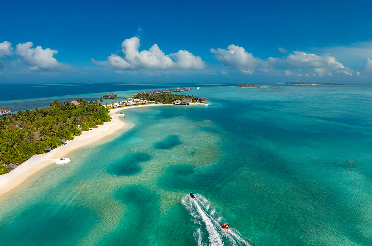 Ariel view over turquoise ocean with an island covered in palm trees to the left of the image, and two speedboats in the water in the foreground