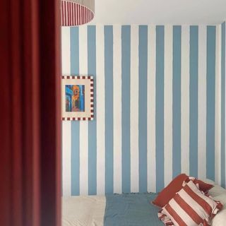 A bedroom with a blue and white striped wallpaper and a striped frame on the wall, a striped cushion on the bed and a striped lampshade