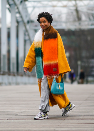 Ellie Delphine wears golden earrings, a multi colored orange and blue striped wool Loewe fringed scarf, an orange wool oversized long coat with flared large sleeves from Mariam Al Sibai, a pullover with colored print, a blue leather bag from Gucci with a golden buckle, gray jogger sportswear pants from Norma Kamali, New Balance sneakers shoes, on January 06, 2021 in Paris, France