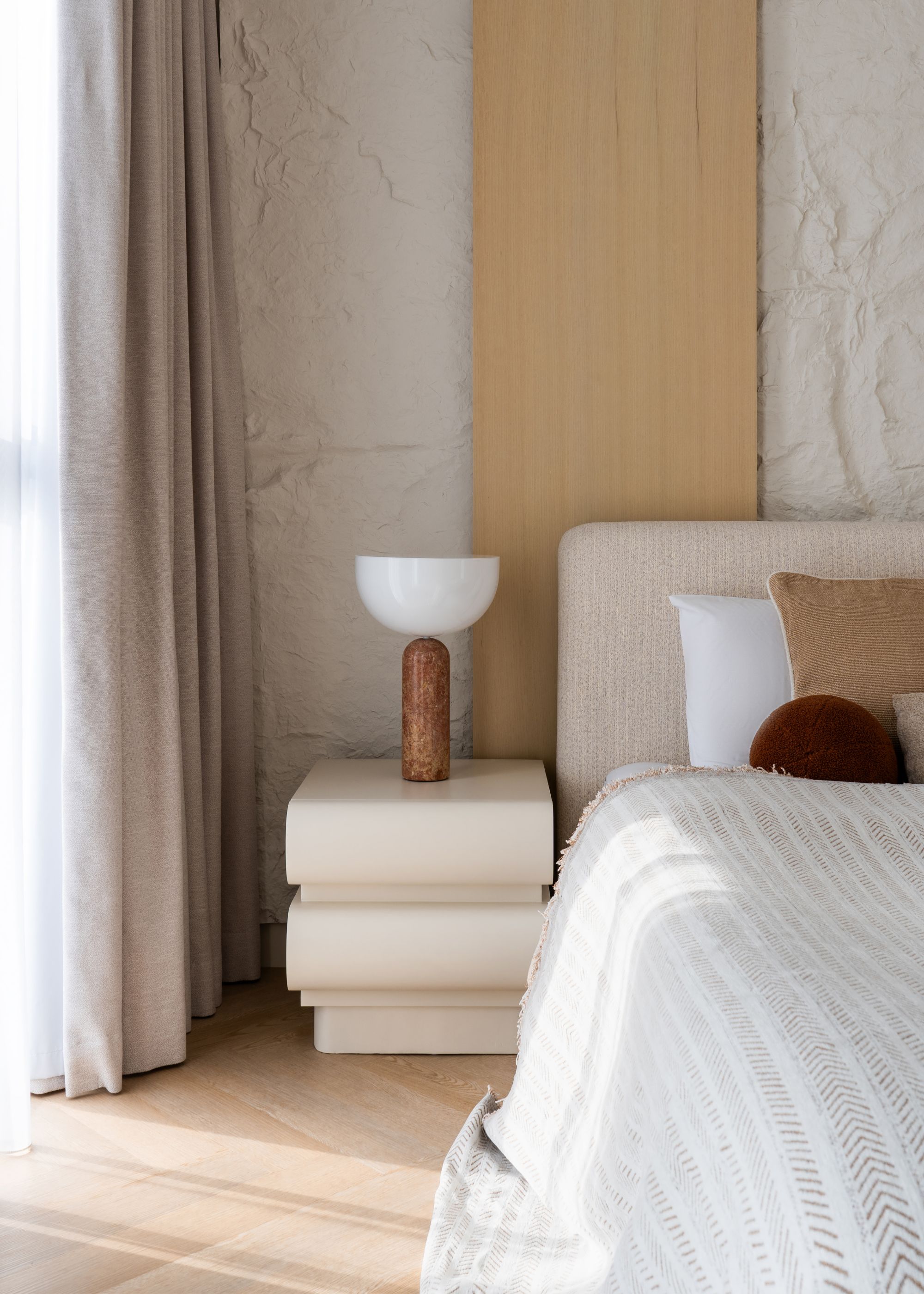 A minimalist bedroom with textured walls, wood accents, a structured side table with a marble base lamp, and a bed dressed in light colors