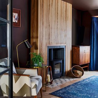 A snug with black Crittall-style framed doors, fluted wood detail around woodburning-stove, blue rug, wooden cabinetry and blue velvet curtain window treatment decor
