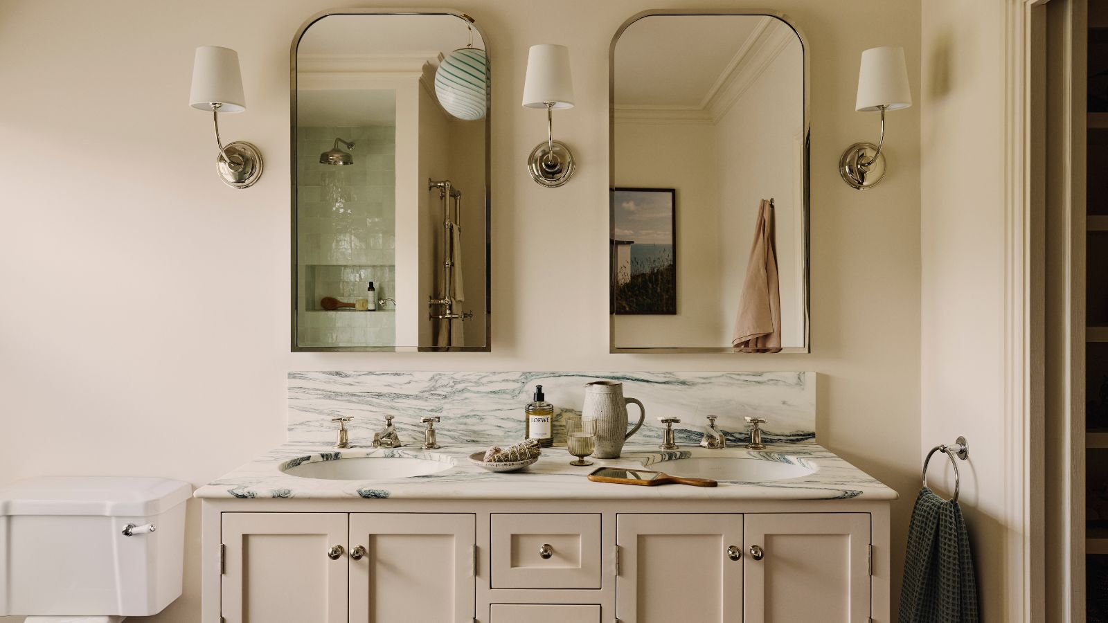 a bathroom with a double vanity and two mirrors above, three wall sconces, decor on the vanity, and a toilet to the left side