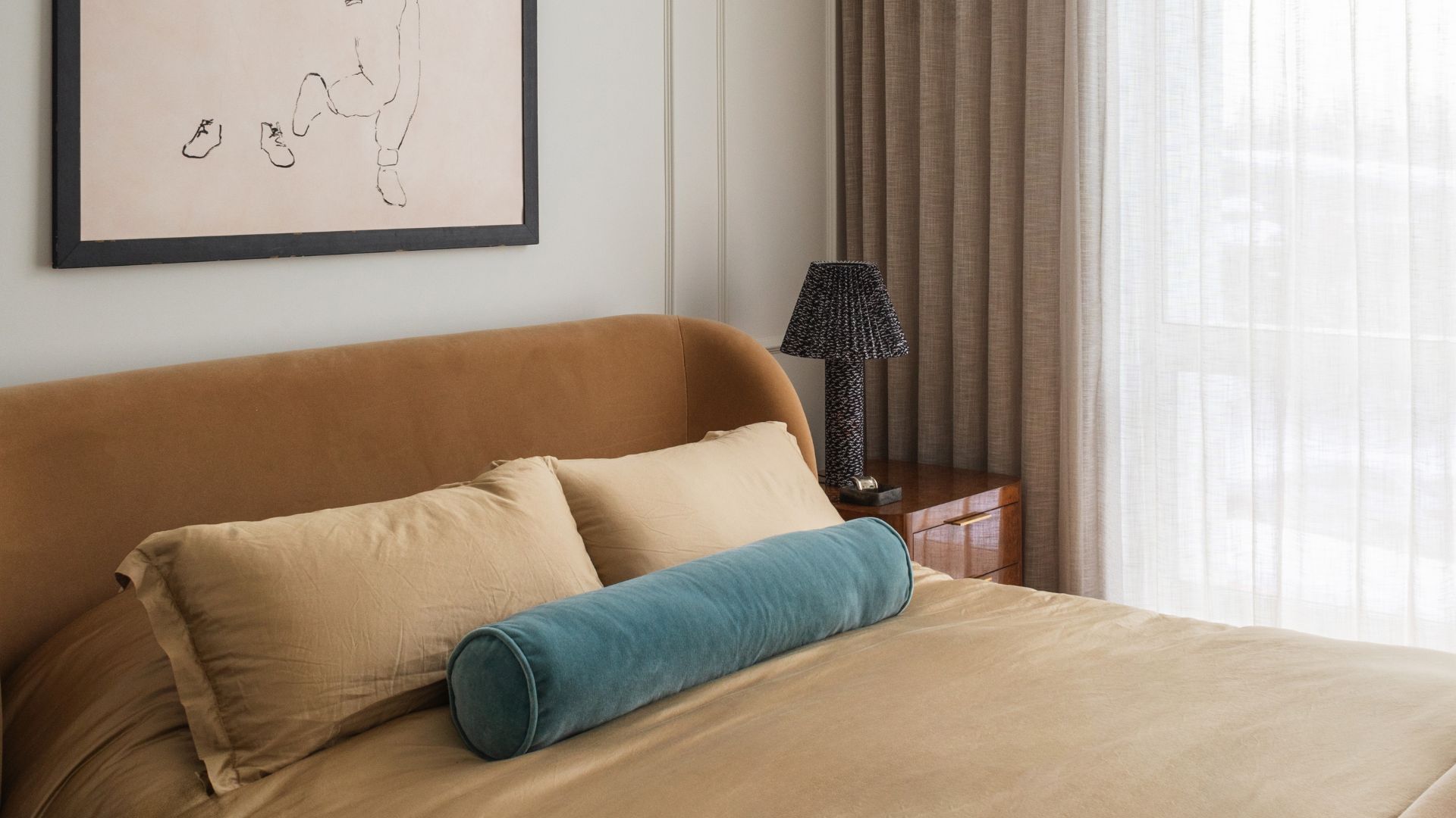 A grounding bedroom with a plush brown bed and upholstered wrap-around headboard, a deep teal bolster pillow, a polished side table, a table lamp and framed artwork