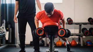 Man performs chest-supported row with dumbbells, another man stands behind him