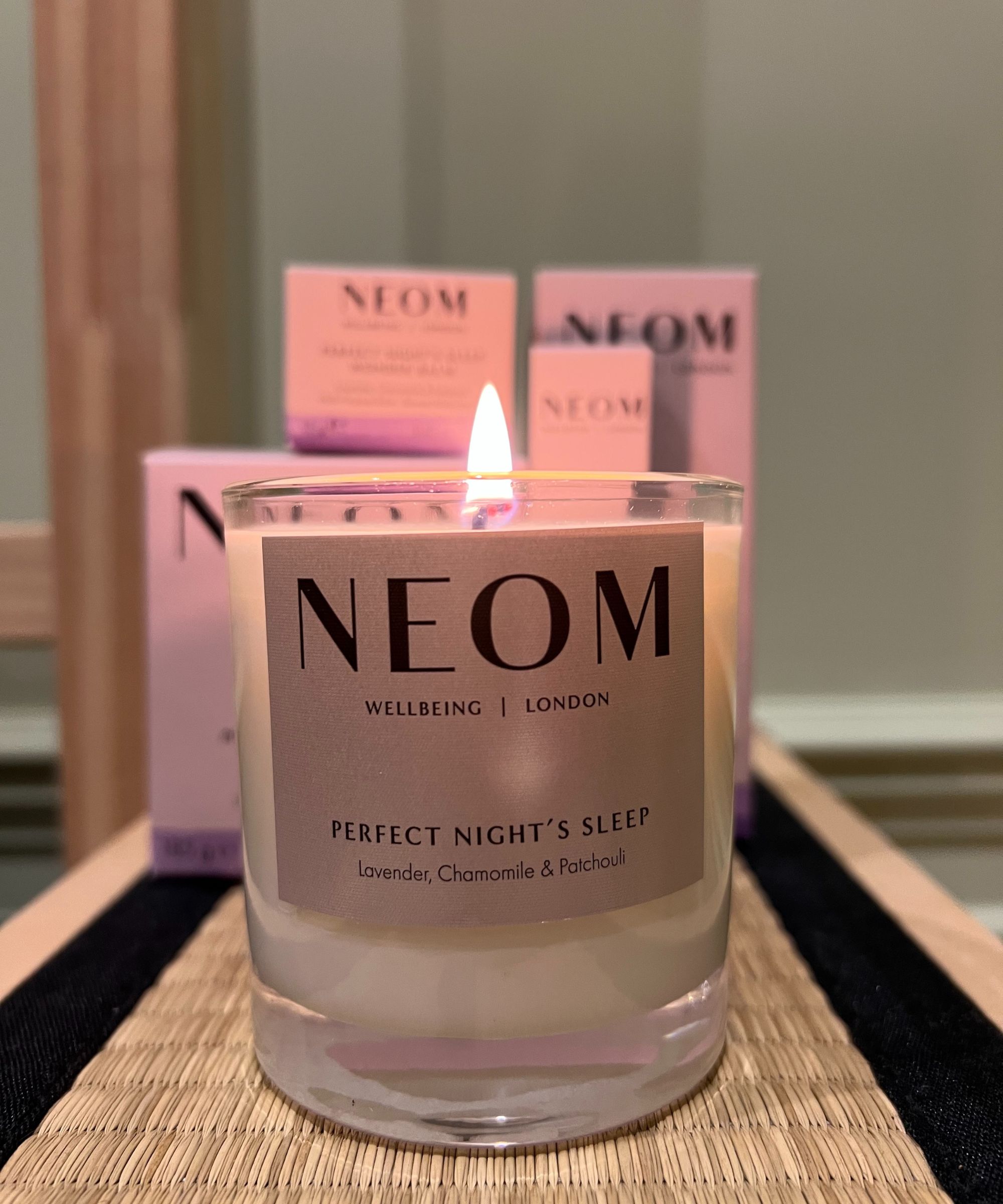 Burning the NEOM Perfect Night's Sleep candle on a tatami mat bedside table, with the whole collection in light purple packaging behind, in front of a wall painted with Farrow &amp;amp; Balls' Green Ground.