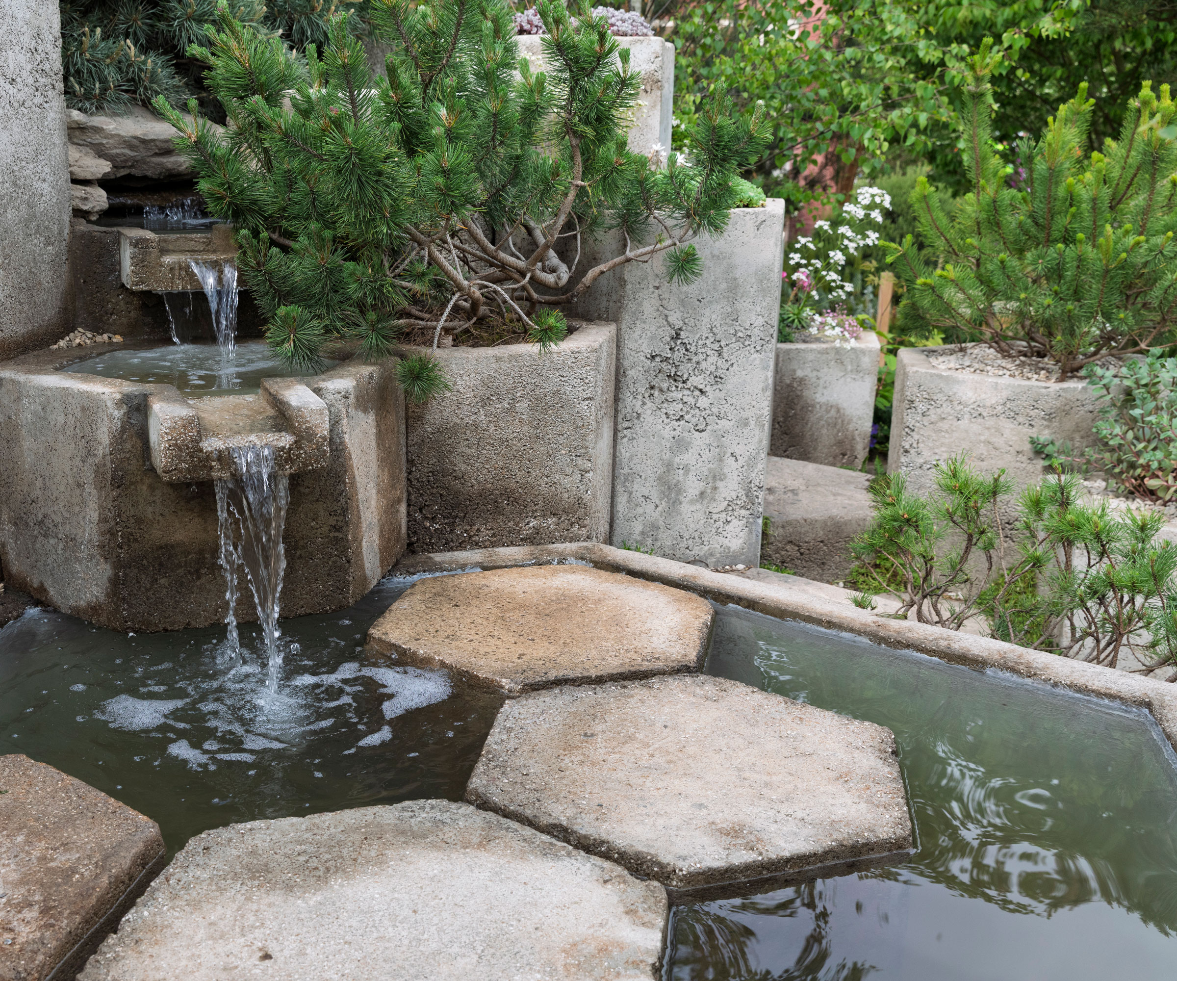 tiered fountain concrete water feature with stepping stone, surrounded by containers of drought tolerant planting including evergreen shrubs