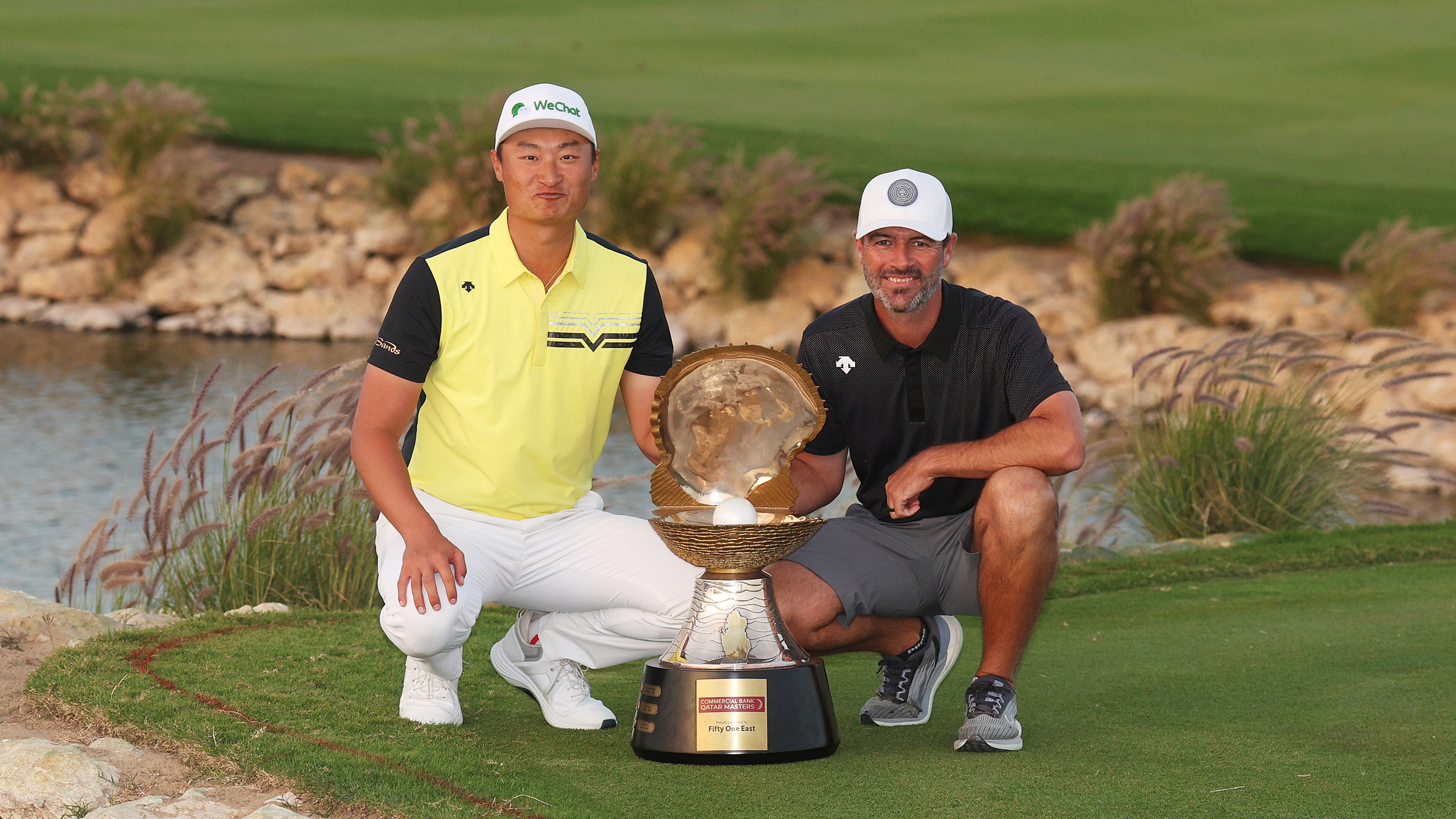 Haotong Li and Jady de Beer crouch down next to the 2025 Qatar Masters trophy