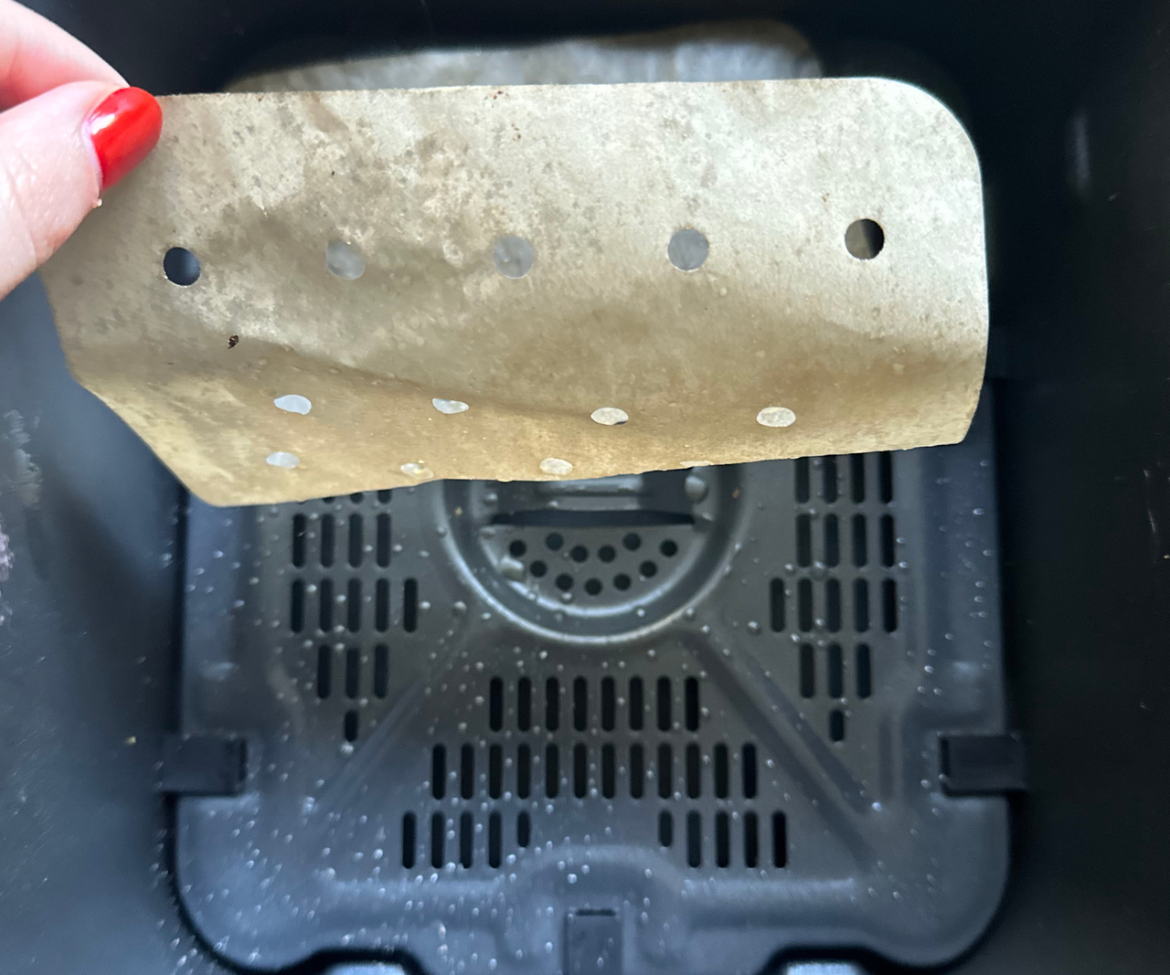 Someone with red painted nails picking up a greasy perforated paper air fryer liner from a black air fryer basket, showing oil on the tray beneath.