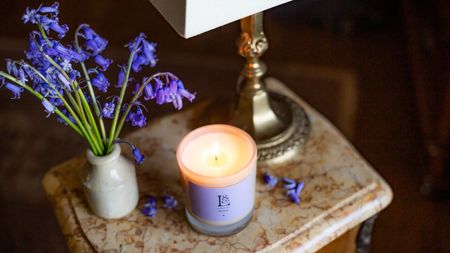 A soy wax candle burning beside a small vase of fresh lavender flowers and an ornate gold based lamp, on a natural stone side table. 