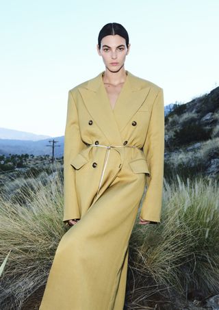 Supermodel Vittoria Ceretti modeling for H&M Studio's spring summer 2024 collection wearing a camel coat belted with a leather cord.