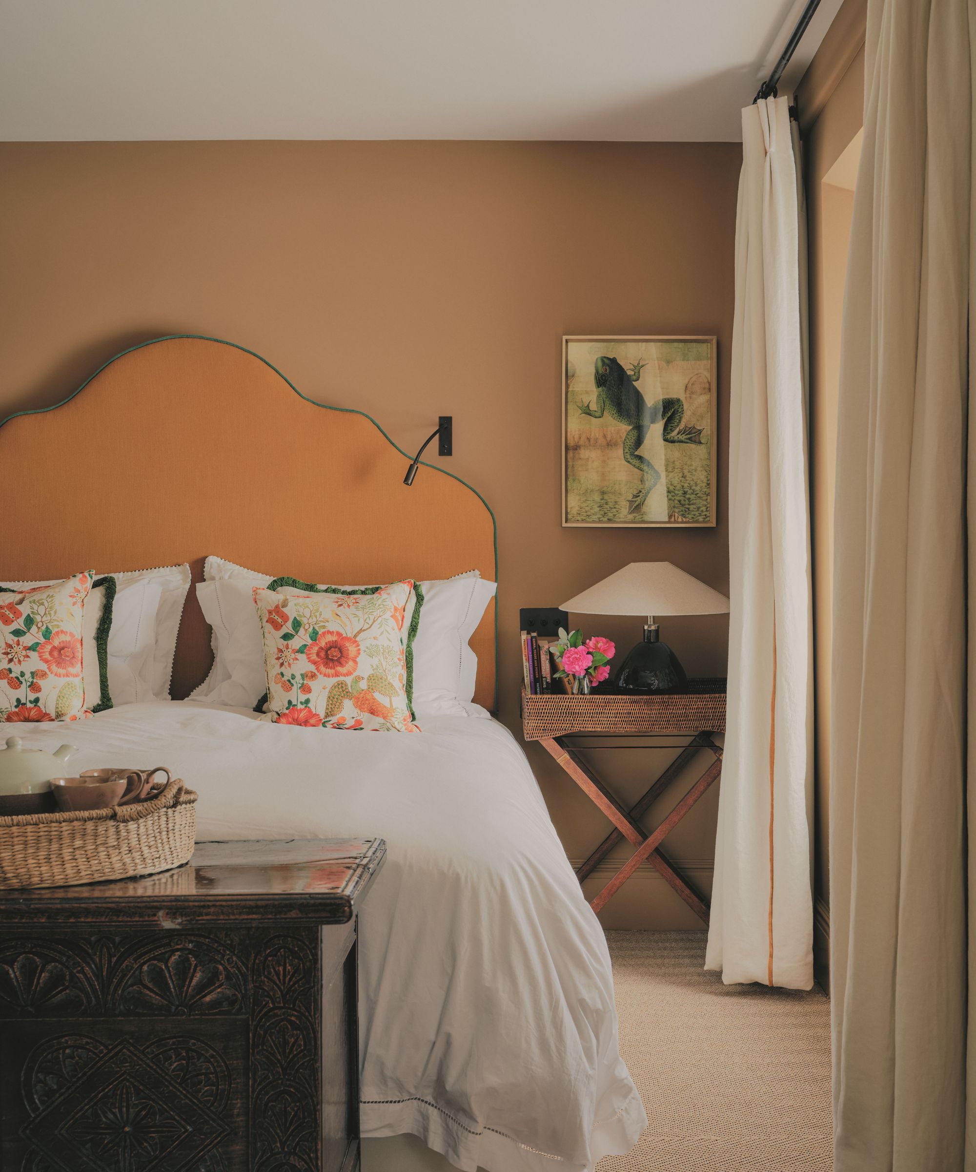 Warm-toned bedroom featuring an orange curved headboard, white bedding with floral accent pillows, a bedside table with a lamp, and framed frog artwork.