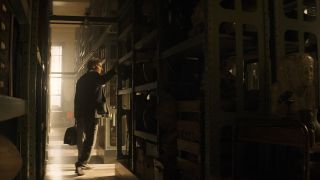 Still from the movie Indiana Jones and the Dial of Destiny. Here we see Indiana Jones wearing a suit and carrying a briefcase as he explores the dark shelves of an archaeological storage room.