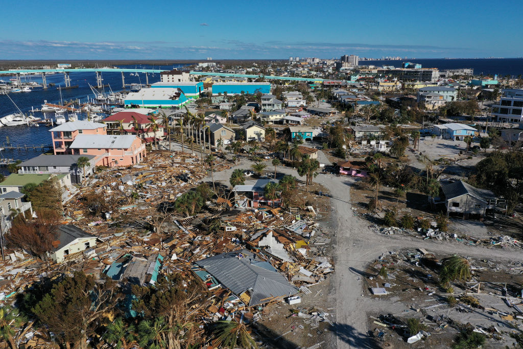 A bird's eye view of a town with the ocean in the background. The town is completely damaged as roofs lie on roadways and buildings are flattened.