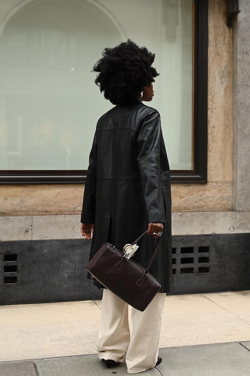 A woman standing outside on the sidewalk turned away from the camera wearing a long black leather duster jacket with off-white wide-leg jeans, black heels, and a brown east-to-west handbag.