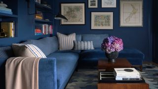 Image of a blue living room with a large blue sectional with a wooden coffee table in front of it. The walls are painted blue with built in bookshelves and art hanging on the wall.