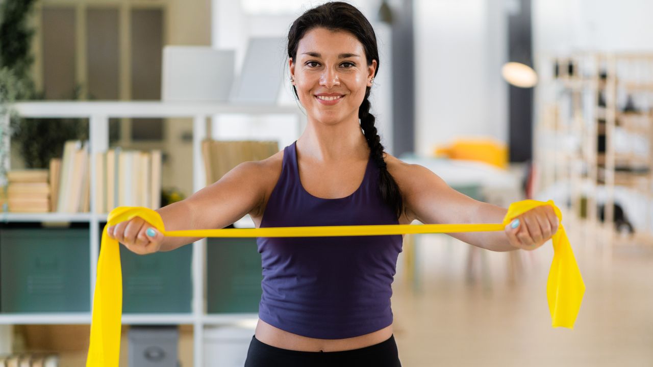 A woman in a sports vest holds a resistance band taut in her hands, with her arms straight. She is smiling. Behind her we see a bookcase, lamp and wooden flooring.