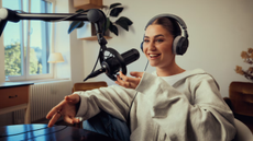 A young woman with dark hair and a light coloured top speaking into a mic while wearing Beyerdynamic DT 270 PRO headphones. She is in a brightly lit room with a window at one side.