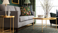 Empire-style glass IKEA coffee table with a gold frame pictured in an opulent living room alongside a coordinating side table, grey sofa, two lamps, and a large sliding glass door.