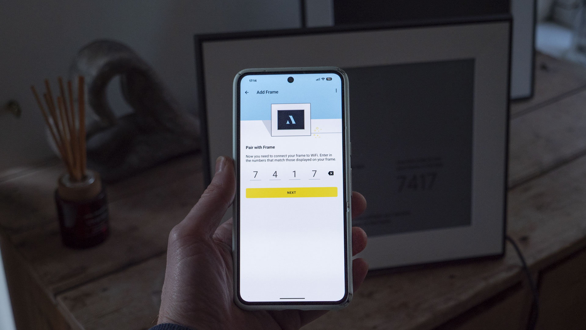 A phone being held, displaying the Aura companion app for digital photo frames, in front of the The Aura Ink 'E Ink' digital photo frame on a sideboard