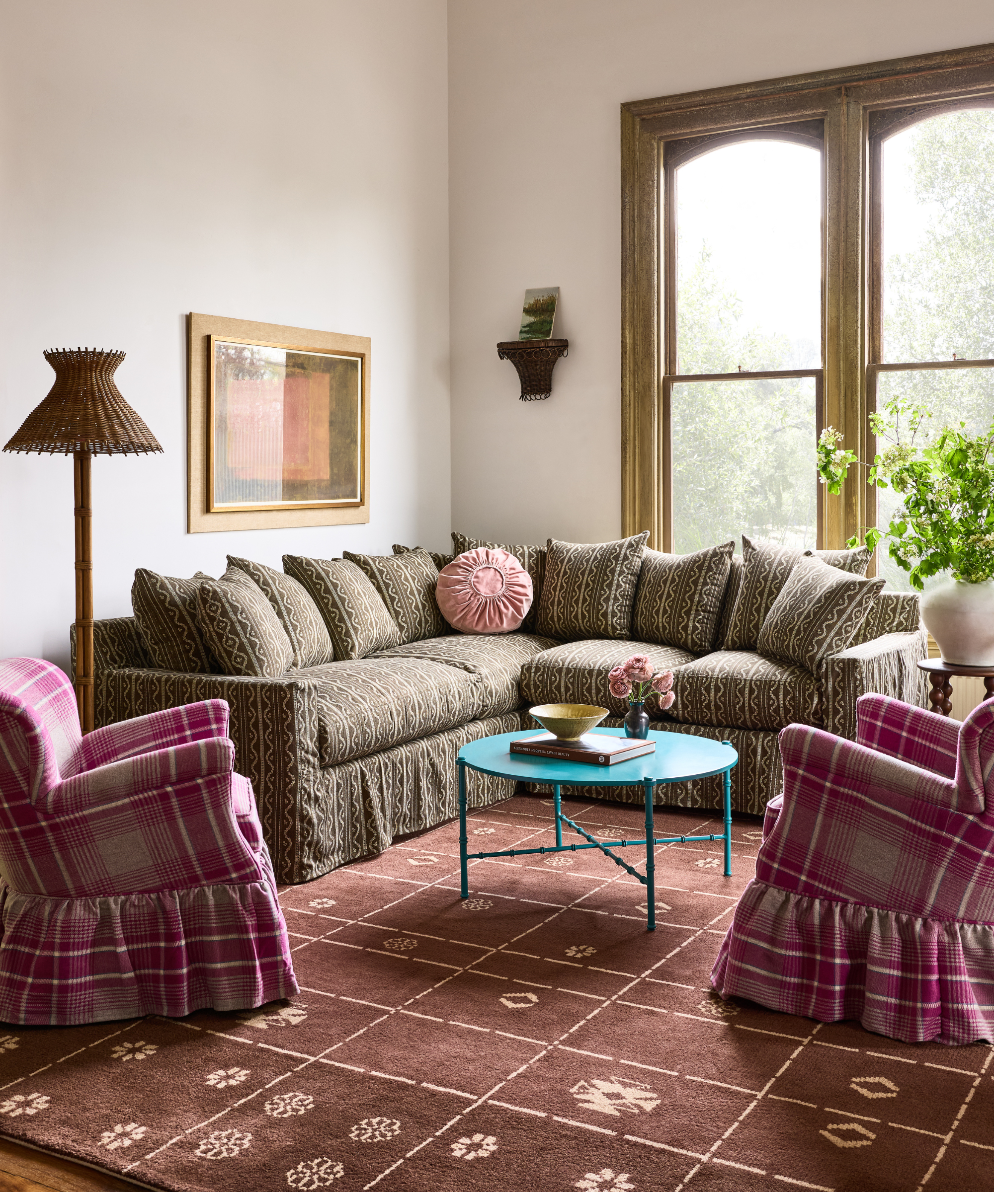 Pattern-drenched living room with large windows, an ikat corner sectional, a pair of plaid skirted accent chairs, chocolate brown geometric rug, and rattan floor lamp.