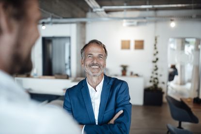 An older man in a blue blazer smiles confidently at the camera. He is talking to a work colleague in the foreground. He looks happy to be at work.