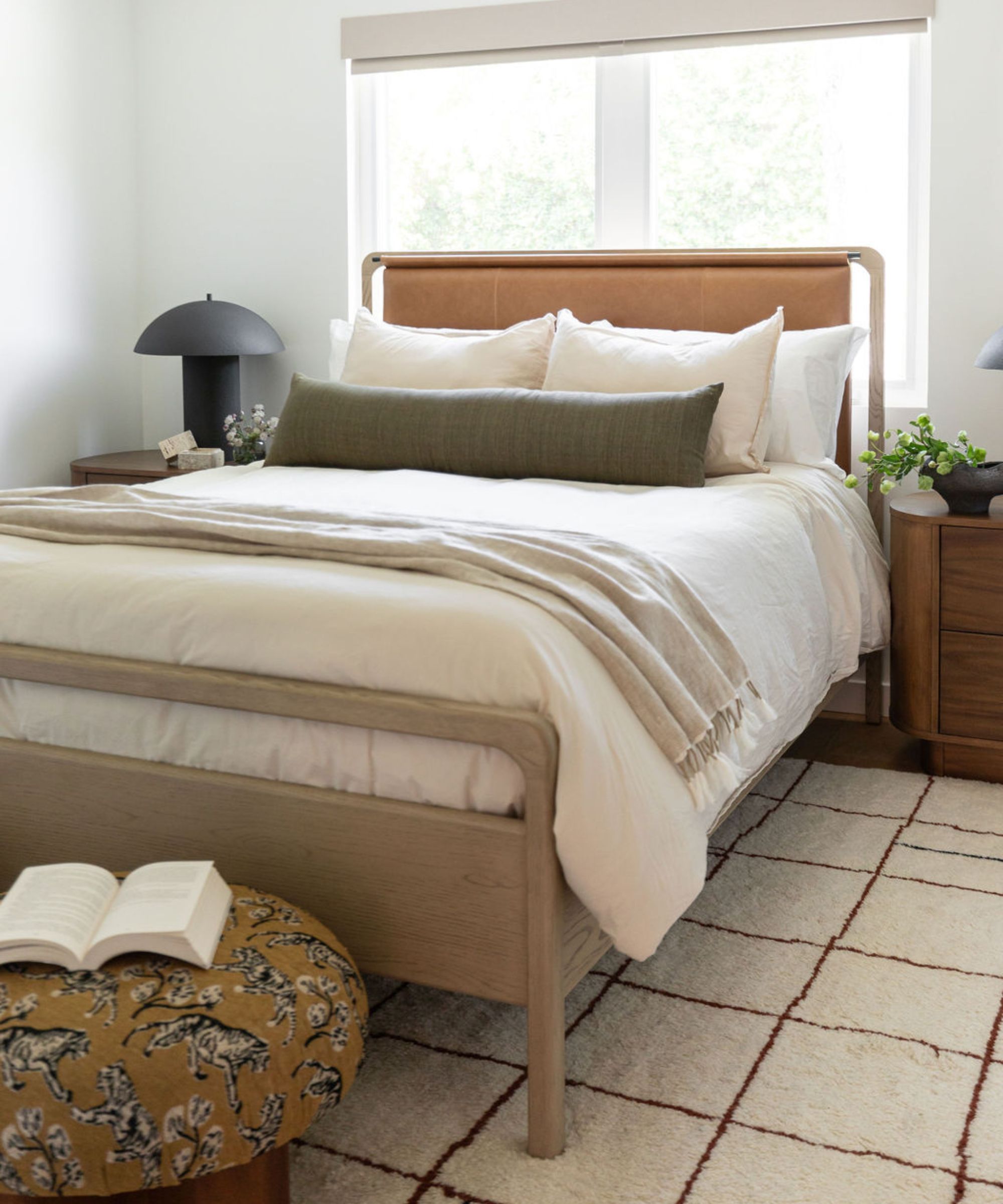 Neutral bedroom with a wooden bed with a leather headboard, linen sheets and a dark brown lumbar pillow, a geometric cream rug, a mustard yellow upholstered ottoman, and a black round table lamp.