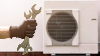 close up of hand wearing black glove holding two spanners with heat pump blurred in background