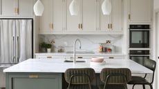 Kitchen island breakfast bar ideas are so chic. Here is a marble kitchen island with a silver sink, sage green base, two green cushioned bar stools, three tulip pendant lights and white cabinets behind it